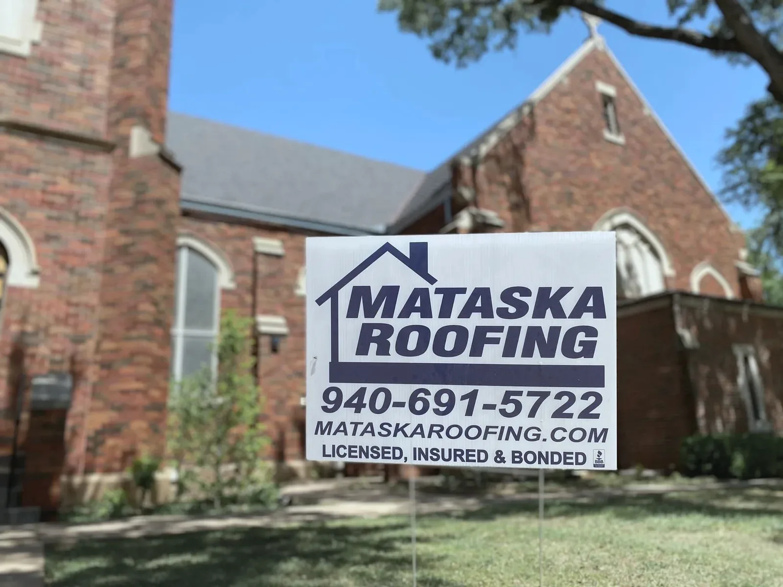 A Mataska Roofing yard sign on the lawn of a red-brick church